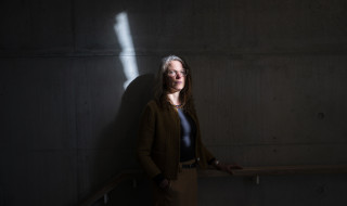 Nolda in de schaduw tegen een muur, een deel van haar gezicht wordt uitgelicht door de zon, ze kijkt ernstig in de lens