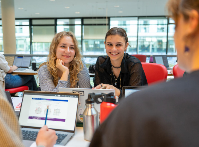 Twee studentes lzitten aan tafel en lachen naar een medestudent 